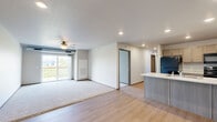 Located in Waukee, Iowa, this three-bedroom, two-bathroom unit offers a thoughtfully arranged single-floor layout designed for comfortable everyday living. The kitchen is a functional highlight, featuring light taupe cabinetry, light-toned countertops with a spacious island, a stainless steel double sink, and black appliances including a refrigerator, range, and over-the-range microwave. Light wood-style flooring runs through the kitchen and entry areas, creating a clean, cohesive aesthetic. The living room is carpeted and features sliding glass doors that open to a private balcony, offering pleasant views of the surrounding greenery and neighborhood landscape. Each of the three bedrooms is carpeted and includes sliding panel closets with a wood-grain finish. The primary bedroom is comfortably sized, while additional bedrooms provide ample space for various uses. Both bathrooms are finished with wood-grain vanity cabinetry, light countertops, wood-style flooring, and full tub-shower combinations. Recessed lighting and neutral wall tones throughout the unit contribute to a bright, clean atmosphere. The overall design is modern and practical, making this a well-appointed unit in a residential Waukee community.