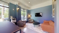 This thoughtfully designed single-floor space functions as a contemporary communal lounge and collaborative work environment. The interior is defined by a cohesive blue-toned color palette applied across the walls, creating a calm and modern atmosphere throughout.

The main lounge area is furnished with a curated mix of seating, including upholstered armchairs, a tan leather sofa, and a sleek oval coffee table, all arranged atop a patterned white area rug. A large flat-screen TV is mounted on the blue accent wall, while a stylish brass floor lamp and globe wall sconces provide warm ambient lighting. Bold artwork, including a prominent crown motif painting, adds an artistic character to the space.

A glass-walled conference or meeting room is accessible via a full-height glass partition and door, offering a semi-private workspace with clear sightlines to the exterior. Adjacent to this is a high-top communal table surrounded by wooden bar-height chairs, positioned beside floor-to-ceiling windows that frame lush outdoor views and flood the area with natural light.

The flooring transitions between polished concrete in the entry and transit zones and soft area rugs in the lounge sections, adding texture and warmth. Recessed ceiling lighting, tray ceiling details, and curated greenery complete the refined, design-forward character of the space.