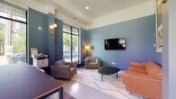 This thoughtfully designed single-floor space functions as a contemporary communal lounge and collaborative work environment. The interior is defined by a cohesive blue-toned color palette applied across the walls, creating a calm and modern atmosphere throughout.

The main lounge area is furnished with a curated mix of seating, including upholstered armchairs, a tan leather sofa, and a sleek oval coffee table, all arranged atop a patterned white area rug. A large flat-screen TV is mounted on the blue accent wall, while a stylish brass floor lamp and globe wall sconces provide warm ambient lighting. Bold artwork, including a prominent crown motif painting, adds an artistic character to the space.

A glass-walled conference or meeting room is accessible via a full-height glass partition and door, offering a semi-private workspace with clear sightlines to the exterior. Adjacent to this is a high-top communal table surrounded by wooden bar-height chairs, positioned beside floor-to-ceiling windows that frame lush outdoor views and flood the area with natural light.

The flooring transitions between polished concrete in the entry and transit zones and soft area rugs in the lounge sections, adding texture and warmth. Recessed ceiling lighting, tray ceiling details, and curated greenery complete the refined, design-forward character of the space.
