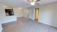 This single-floor residence offers a straightforward and functional layout designed for comfortable everyday living. The main living area is a standout feature, showcasing light-toned hardwood-style flooring that extends throughout the space, creating a bright and cohesive aesthetic. Neutral, warm-beige walls complement the light flooring and contribute to an airy, welcoming atmosphere. A ceiling fan with integrated lighting is centrally mounted, providing both ventilation and ambient illumination to the living space. The open layout allows for flexible furniture arrangement and connects seamlessly to adjacent areas. A partial view of the kitchen is visible along one side, hinting at a practical and accessible kitchen design with dark appliances and a light countertop peninsula or bar. Multiple doorways lead to additional rooms, suggesting a well-organized floor plan with clearly defined private and common spaces. White interior doors and white baseboards throughout add a crisp, clean contrast to the warm wall tones. The overall design is move-in ready with a minimalist, modern-neutral character that suits a wide range of uses and personal styles.