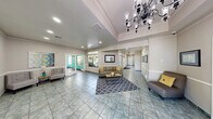 Located at 4640 Vegas Valley Dr in Las Vegas, NV, this single-floor community clubhouse offers a thoughtfully designed space combining social comfort with fitness amenities. The spacious hallway and lobby area immediately impress with elevated ceilings adorned with decorative crown molding and a striking ornate chandelier as a central focal point. Large-format slate-toned tile flooring runs throughout, lending a clean, polished aesthetic to the entire common area. Natural light pours in through multiple sets of French doors and windows, creating a bright and inviting atmosphere. The lounge area is furnished with a neutral-toned sofa, accent chairs with yellow cushions, and a patterned area rug, offering a comfortable gathering space. Framed artwork and wall sconces add tasteful decorative touches, while additional French doors lead to adjacent rooms, including a windowed office or leasing area. The exercise room is vibrantly appointed with bold yellow and teal accent walls, hardwood-style flooring, and floor-to-ceiling mirrors that amplify the sense of space. It is equipped with a multi-station cable machine, a treadmill, free weights, and a ceiling fan, providing a well-rounded fitness environment with views of outdoor greenery through large windows.