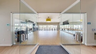 Located at 8920 W Russell Rd in Las Vegas, NV, this expansive fitness and wellness facility offers a comprehensive suite of spaces designed to support a full range of health and exercise activities. The main gym floor is a spacious, light-filled area featuring high ceilings, large floor-to-ceiling windows that bring in abundant natural light and outdoor views of the surrounding landscape, and vibrant motivational wall murals. It is outfitted with an extensive array of cardio machines including treadmills and stair climbers, a full free-weight section with multi-tiered dumbbell racks, cable machines, benches, and functional training equipment. Striking yellow pendant lights and ceiling fans enhance the bright, energetic ambiance. A dedicated Pilates studio houses multiple reformers and tower units arranged on light hardwood flooring beneath soaring ceilings. Adjacent to it, a movement and yoga studio features a full wall of mirrors, large windows overlooking the exterior, and open floor space suited for mat-based classes and group training. Two serene treatment rooms provide a calm, spa-like setting with soft neutral tones, warm accent lighting, massage tables, and tasteful artwork. Hallways connect each area smoothly, creating an efficient, professional layout throughout this well-appointed wellness center.