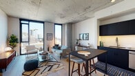 This contemporary urban residence showcases a striking industrial-modern aesthetic defined by exposed concrete ceilings and structural columns that contrast beautifully with crisp white walls and light hardwood flooring throughout. The open-plan living and dining area is the heart of the space, anchored by dramatic floor-to-ceiling windows that frame sweeping city skyline views and flood the interior with abundant natural light. A high-top dining table with bar seating transitions seamlessly into the living area, which features a comfortable lounge arrangement set atop a patterned area rug. The kitchen is outfitted with matte navy cabinetry, white subway tile backsplash, stainless steel appliances including a full-size refrigerator, microwave, and dishwasher, complemented by under-cabinet lighting. The bedroom is a serene retreat with its own floor-to-ceiling window overlooking the cityscape, exposed concrete column, and ample space for a full furnishing arrangement. The bathroom presents a clean, graphic design with black hexagonal mosaic floor tiles, white grid-pattern shower tiles, a glass-enclosed walk-in shower, and a sliding closet door providing direct access to built-in storage. Together, these elements create a cohesive, sophisticated living environment with an unmistakable urban edge.