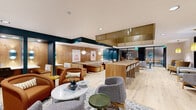 Located at 2650 University Ave SE in Minneapolis, MN, this thoughtfully designed single-floor amenity space offers a dynamic blend of collaborative and private environments. The open-plan main area is anchored by light hardwood flooring and a striking teal and warm wood color palette, creating an inviting yet energized atmosphere throughout. A long communal bar-height counter with white upholstered seating serves as a central hub, complemented by cushioned booth seating along wood-paneled walls adorned with curated artwork. Multiple lounge zones are furnished with a curated mix of rust-orange armchairs, houndstooth accent chairs, and terrazzo-topped tables, offering flexible seating for both focused work and casual gatherings. Recessed and pendant lighting, including globe and linear drop fixtures, provide warm, layered illumination across the space. A dedicated quiet lounge features plush sectional and accent seating with wall-mounted display screens, ideal for more relaxed or private use. Built-in desk nooks with teal accent lighting offer semi-private individual workstations, while glass-enclosed phone booths and a private office room support focused tasks and confidential conversations. A separate lounge area with yellow pendant lighting and a pegboard-textured accent wall adds a distinct, vibrant character to one end of the floor, rounding out a well-appointed, multifunctional space.