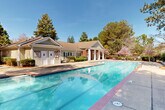 This outdoor amenity space centers around a generously sized rectangular swimming pool with crystal-clear turquoise water and classic brick-edged coping. The pool is accessible via built-in metal handrails and is clearly marked with depth indicators, reflecting a strong emphasis on safety alongside aesthetics. A wide concrete pool deck surrounds the water on multiple sides, providing ample room for lounging and circulation. Rows of sling-style lounge chairs line the perimeter, offering comfortable spots for sunbathing, while umbrella-shaded tables and chairs are available for those seeking shade. A paved patio area with additional chaise lounges sits adjacent to the pool, framed by tall, mature pine trees and leafy hardwoods that provide a natural, park-like backdrop. The entire area is enclosed by a black wrought iron fence with manicured shrubbery and planted beds along the border, lending both security and a polished appearance. A pool house or amenity building with white exterior siding, columns, and a pitched roof is situated at one end, housing restrooms and pool-side facilities. The open-sky setting allows abundant natural light throughout the day, creating a bright and inviting atmosphere ideal for relaxation and recreation.