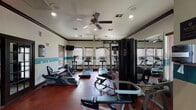 This fitness center in Houston, TX offers a comprehensive workout environment designed to support a wide range of exercise routines. The space is spread across a single floor and is divided into distinct functional zones, each catering to different training needs.

The main cardio area is generously sized and lined with multiple treadmills, elliptical trainers, and a recumbent bike, all positioned in front of a row of windows fitted with wood-slatted blinds that allow natural light to fill the room. Ceiling fans overhead provide additional air circulation, complementing the recessed lighting throughout. Wall-mounted flat-screen televisions are positioned for easy viewing during workouts.

A dedicated strength training section houses a multi-station cable and weight machine alongside free-weight benches, while a mirrored wall amplifies the sense of space and allows users to monitor their form. A separate area features a full dumbbell rack with a wide range of weights, suspension training straps, and a heavy punching bag, catering to functional and combat-style fitness routines.

The interior is finished with a bold teal accent stripe running along the walls, dark crown molding, and rich red rubber flooring throughout, creating a vibrant and energetic atmosphere. French-style glass doors provide entry to the space, adding a refined architectural touch to this practical and fully equipped fitness facility.