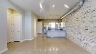 This two-floor space masterfully combines industrial character with modern design sensibilities. The open-concept main level immediately draws attention with its striking exposed brick accent wall, which adds warmth and texture against the clean, neutral-toned walls and smooth white ceilings. Polished large-format tile flooring spans the living areas, reflecting light and enhancing the sense of spaciousness throughout.

The kitchen is a standout feature, designed with sleek, light wood cabinetry and a generous island that serves as both a prep and gathering space. Stone countertops add a refined touch, while a trio of pendant lights hang above the island, providing focused task lighting with a contemporary aesthetic. A stainless steel refrigerator with an integrated dispenser anchors one end of the kitchen, complemented by white upper cabinetry and a window above the sink that brings in natural light. Recessed ceiling lighting throughout ensures the space is well-illuminated at all hours.

Exposed ductwork along the upper brick wall adds an authentic industrial accent, seamlessly integrated into the overall design. A hallway leads to additional rooms, offering functional separation within the open layout. With its thoughtful blend of raw materials and modern finishes, this space presents a polished yet characterful environment across both floors.