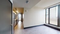 This single-floor unit showcases a contemporary design with clean lines and a neutral color palette throughout. The open layout connects the living area to the galley-style kitchen, which is equipped with stainless steel appliances, including a refrigerator, range, and built-in microwave. White cabinetry with sleek hardware, light quartz countertops, and a tiled backsplash give the kitchen a polished, modern aesthetic. Track lighting runs along the ceiling, providing functional illumination across the workspace.

Wood-look plank flooring extends throughout the main living areas, complemented by dark baseboards that add a subtle contrast to the light walls. Multiple large, black-framed windows are positioned throughout the unit, drawing in generous amounts of natural light and offering open urban views. The spacious living area benefits from multiple window exposures, enhancing the sense of openness.

The bedroom features additional large windows with exterior views and is outfitted with a ceiling fan. The bathroom is finished with white square tile from floor to ceiling, and includes a soaking tub positioned beneath a window that frames an expansive city and waterfront view, adding a distinctive architectural detail to the space.