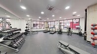 This modern fitness center offers a generously sized, single-floor workout space thoughtfully designed to accommodate a wide range of exercise needs. The layout is open and efficiently organized, allowing for comfortable movement between equipment zones. Large, black-framed windows line the exterior walls, providing abundant natural light and street-level views of the surrounding neighborhood, creating an energizing atmosphere for exercise. Recessed circular lighting complements the natural light, ensuring the space is well-illuminated throughout. The flooring consists of durable rubber gym matting in a speckled gray finish, providing both safety and stability during workouts. Exposed concrete columns add an industrial architectural character to the otherwise clean, white-walled interior. Full-length wall mirrors span a significant portion of the space, visually expanding the room while allowing users to monitor their form. Equipment includes multiple treadmills, an elliptical trainer, a stationary bike, a cable weight machine, a multi-tiered dumbbell rack with a full range of weights, kettlebells, medicine balls, adjustable benches, and a barbell weight rack. A ceiling-mounted HVAC cassette unit ensures climate control comfort. The overall ambiance is clean, bright, and functional, reflecting a professional fitness facility environment.