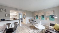 This single-floor residence offers a thoughtfully designed open-concept layout that seamlessly connects the living, dining, and kitchen areas. Light wood-style flooring runs throughout the main living spaces, complementing the neutral gray walls and textured ceilings, while creating a cohesive and inviting atmosphere.

The living area is bright and welcoming, featuring a large window that frames views of the surrounding greenery and allows natural light to fill the room. Bold abstract artwork in teal and blush tones adds a contemporary touch to the space.

Adjacent to the living area, the dining space accommodates a glass-top table with seating for four, flowing naturally from the kitchen. The kitchen features a peninsula with a dark countertop and bar stool seating, white cabinetry, subway tile backsplash, stainless steel appliances, and a window that brings in additional natural light.

The bedroom is carpeted for comfort and includes a large window with outdoor views, a closet, and a serene, restful ambiance. The bathroom is finished with a dark stone-style countertop, a wide vanity mirror with white framing, wood-style flooring, and brushed nickel fixtures, giving it a clean and modern feel throughout.