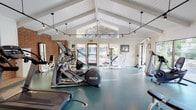 This fitness center is housed in a thoughtfully designed single-story structure distinguished by its striking vaulted, beam-lined ceiling with white shiplap paneling that lends an open, airy quality to the interior. The soaring roofline creates generous vertical space throughout, enhancing the sense of openness and comfort during workouts.

The facility is generously sized, accommodating a broad range of exercise equipment with ample room to move freely between stations. Cardio equipment includes multiple treadmills, elliptical trainers, and stationary bikes from recognized brands such as Cybex and Precor. Strength training is supported by a dedicated cable machine and a variety of weight equipment positioned throughout the space.

Natural light flows in abundantly through large windows and glass doors on multiple sides of the building, offering pleasant views of surrounding greenery and landscaping. This connection to the outdoors enhances the overall atmosphere. Exposed brick accent walls provide a warm, textural contrast against the crisp white ceiling and light-toned walls.

Underfoot, durable teal rubberized flooring covers the entire workout area, offering both cushioning and easy maintenance. Track-style pendant lighting supplements natural light, ensuring consistent illumination. A mounted flat-screen television is also present, adding convenience for users during extended workout sessions.
