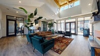 Located at 17014 Fairmount Way in Charlotte, NC, this single-floor clubhouse leasing office presents a welcoming and upscale environment designed to impress. The space is anchored by a dramatic vaulted ceiling with a striking wood-paneled skylight detail that draws the eye upward, while floor-to-ceiling black-framed windows and glass doors flood the interior with natural light and offer views of the surrounding community.

The main lounge area is thoughtfully appointed with a rich cognac leather sofa, deep teal velvet accent chairs, and a live-edge wood coffee table, all grounded by a vibrant jewel-toned area rug. Towering tropical plants punctuate the space, adding lush greenery and a sense of organic vitality. A built-in feature wall on one side houses an electric fireplace beneath a wall-mounted television, flanked by open shelving with warm wood accents, creating a cozy yet contemporary focal point.

Wood-look tile flooring flows throughout, complementing the neutral walls and modern finishes. Multiple entryways with glass door panels connect to exterior views, including a covered patio and pool area. Tufted bench seating near the entry points provides a practical yet refined transition into the space. The overall ambiance is modern, airy, and inviting — balancing sophisticated design with a comfortable, community-oriented atmosphere.