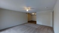 This single-floor space offers a clean, open layout with a straightforward and functional design. The main living area is finished with wall-to-wall carpeting in a neutral tone, providing a comfortable and warm underfoot feel throughout. Smooth, light-colored walls complement the understated aesthetic, giving the space a blank canvas quality well-suited for personalization. A ceiling fan with a built-in light fixture is centrally mounted, ensuring both air circulation and ambient lighting. The textured popcorn ceiling adds a classic touch overhead. Toward the rear of the space, the flooring transitions to a darker hardwood or laminate surface, visually distinguishing a separate functional zone — likely a dining or utility area. Flush-mounted ceiling vents are positioned throughout, indicating a central HVAC system for year-round climate control. Subtle baseboard trim runs along the perimeter, adding a clean finish to the walls. The overall ambiance is calm and neutral, with an open feel that allows for flexible furniture arrangements and adaptable use of the available space.