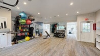 This fitness center offers a thoughtfully designed single-floor layout that accommodates a full range of workout activities. The space is divided into two functional zones, seamlessly connected to support both cardio and strength training needs. Durable, patterned carpet flooring runs throughout, providing comfort underfoot while maintaining a clean, polished aesthetic.

The main workout area is generously sized, featuring large floor-to-ceiling mirrors that visually expand the space and allow for form monitoring during exercise. Equipment includes treadmills, an elliptical trainer, a stationary bike, and a multi-function power tower for bodyweight training. A well-stocked free weights section houses a full rack of dumbbells and kettlebells alongside an adjustable workout bench. A dedicated storage rack holds an assortment of medicine balls, stability balls, and resistance training accessories, including TRX suspension straps.

Natural light fills the space through multiple windows along the walls, creating a bright and energizing atmosphere, while recessed ceiling lighting ensures consistent illumination throughout. An accent wall featuring a scenic mural adds a distinctive design element to the strength training area.

The entry zone includes practical amenities such as open cubbies for personal storage, a built-in water fountain, a fire safety station, and posted fitness rules, ensuring a safe and organized environment for all users.