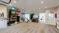 This fitness center offers a thoughtfully designed single-floor layout that accommodates a full range of workout activities. The space is divided into two functional zones, seamlessly connected to support both cardio and strength training needs. Durable, patterned carpet flooring runs throughout, providing comfort underfoot while maintaining a clean, polished aesthetic.

The main workout area is generously sized, featuring large floor-to-ceiling mirrors that visually expand the space and allow for form monitoring during exercise. Equipment includes treadmills, an elliptical trainer, a stationary bike, and a multi-function power tower for bodyweight training. A well-stocked free weights section houses a full rack of dumbbells and kettlebells alongside an adjustable workout bench. A dedicated storage rack holds an assortment of medicine balls, stability balls, and resistance training accessories, including TRX suspension straps.

Natural light fills the space through multiple windows along the walls, creating a bright and energizing atmosphere, while recessed ceiling lighting ensures consistent illumination throughout. An accent wall featuring a scenic mural adds a distinctive design element to the strength training area.

The entry zone includes practical amenities such as open cubbies for personal storage, a built-in water fountain, a fire safety station, and posted fitness rules, ensuring a safe and organized environment for all users.