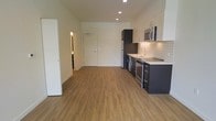 This single-floor space offers a thoughtfully designed open-concept layout that seamlessly integrates the living area and kitchen into one cohesive environment. The warm, medium-toned hardwood-style flooring runs throughout, lending a welcoming and cohesive aesthetic to the space.

The kitchen is a standout feature, showcasing a sleek, modern design with dark cabinetry contrasted by crisp white upper cabinets and a classic white subway tile backsplash. Stainless steel appliances — including a refrigerator, stove, and over-the-range microwave — are complemented by a clean white countertop and a contemporary sink with a modern faucet. Recessed lighting illuminates the kitchen area effectively, highlighting its clean lines and quality finishes.

The open living area provides generous room for flexible furnishing arrangements. Bright white walls and recessed ceiling lights contribute to an airy, well-lit atmosphere throughout. A hallway visible from the main living space leads to additional rooms, suggesting a functional and efficient floor plan. The overall ambiance is clean, contemporary, and move-in ready, making it an appealing space for those who appreciate modern, minimalist design with practical everyday functionality.