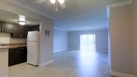 This single-story space presents an open and airy layout characterized by clean lines and a neutral color palette. The main living area features light-toned tile flooring that reflects natural light, contributing to a bright and welcoming atmosphere. Crown molding runs along the perimeter of the ceiling, adding a subtle architectural detail that elevates the overall finish of the space.

A ceiling fan is centrally mounted, providing both functional air circulation and a modest decorative element. At the far end of the living area, a sliding glass door fitted with vertical blinds serves as the primary source of natural light and provides access to an outdoor area, offering views beyond the interior.

Adjacent to the main living space, a partial wall separates the kitchen area, where a white refrigerator is visible alongside dark cabinetry, suggesting a functional kitchen setup. An electrical panel is mounted on the wall near the kitchen entrance, a practical feature discretely integrated into the layout. The overall design is straightforward and functional, with consistent finishes throughout that create a cohesive and well-maintained environment.