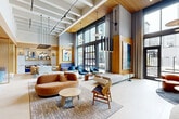 This thoughtfully designed amenity lounge offers a generous, open-plan layout that makes a strong architectural impression from the moment you enter. Soaring double-height ceilings with exposed structural beams and warm wood-paneled accents create a striking visual contrast against the light-toned tile flooring below. Floor-to-ceiling windows and glass-panel doors flood the interior with abundant natural light while framing direct views of and access to an outdoor courtyard and pool area beyond.

The main lounge area is anchored by a curated collection of contemporary furnishings, including a cognac leather sofa, sculptural wooden coffee tables, and an assortment of accent chairs in rich blues and earthy tones. A standout built-in fireplace feature wall, clad in smooth concrete and topped with a wall-mounted display, serves as a dramatic focal point and is complemented by organic greenery and designer wall sconces.

A semi-private seating nook with a distinctive oval glass partition offers an intimate retreat within the larger space, furnished with plush velvet banquette seating and lounge chairs. Adjacent to the main lounge, a bright counter-height bar area provides an additional social gathering space. Curated large-scale artwork is displayed throughout, reinforcing the space's refined, gallery-like aesthetic. The overall ambiance is modern, warm, and inviting.