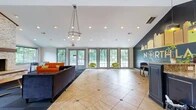 Located at 2905 Cottage Pl in Greensboro, NC, this single-floor community clubhouse for North Lakes offers a thoughtfully designed, open-plan gathering space. The layout transitions seamlessly between distinct functional zones, all united by a combination of large-format tile flooring with diamond accent inlays and plush dark carpet.

The main lounge area is anchored by a striking floor-to-ceiling stacked-stone fireplace with a wood mantel, complemented by vaulted ceilings and recessed lighting. A stylish hoop chandelier adds a modern focal point overhead. Vibrant seating — including an orange sofa and bold green accent chairs — creates an inviting, contemporary atmosphere. A wall-mounted television is positioned nearby for added convenience.

A wall of tall, grid-pane windows and glass doors floods the space with natural light and provides views of the lush, tree-lined outdoor surroundings. A foosball table adds a recreational element to the room.

The kitchen area is outfitted with dark cabinetry, granite countertops, stainless steel appliances, and a spacious bar-height island with metal stools — ideal for casual dining or socializing. Pendant lighting and live greenery add warmth to this zone.

The space is further distinguished by a bold "North Lakes" branded accent wall in deep charcoal with a vibrant yellow cityscape mural, reinforcing the community's identity throughout.