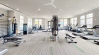 Located at Crest Preserve in Bonita Springs, Florida, this fitness center offers a generously sized, single-floor exercise room designed to support a comprehensive workout experience. The space is bright and airy, with large windows lining multiple walls that allow abundant natural light to fill the room and provide views of the surrounding outdoor environment. Overhead ceiling fans complement the recessed lighting, ensuring a comfortable atmosphere throughout.

The exercise room is well-organized and thoughtfully outfitted with a diverse selection of equipment. Cardio machines — including treadmills, ellipticals, and stationary bikes — are positioned to take advantage of the natural light from the windows. A multi-station cable machine anchors the strength training area, accompanied by a variety of benches, free weights, and resistance machines to support full-body workouts. A dedicated wall-mounted storage rack holds medicine balls, dumbbells, and other accessories, catering to functional and dynamic training routines.

The flooring features neutral-toned, striped carpet with subtle accent details, contributing to the clean, modern aesthetic of the space. Soft gray walls and a tray ceiling detail add architectural refinement, while multiple entry points provide convenient access. Overall, this fitness center delivers a well-rounded, functional, and inviting environment for residents seeking an on-site exercise facility.