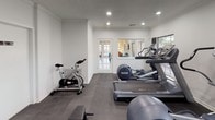 This single-floor fitness center is thoughtfully designed to accommodate a full range of workout activities. The space is divided into functional zones, each serving a distinct fitness purpose. The cardio area is outfitted with multiple treadmills, elliptical trainers, and a stationary spin bike, several of which are equipped with individual monitor screens for entertainment during workouts. Natural light filters in through multiple windows, offering pleasant outdoor views and creating a bright, energizing atmosphere alongside recessed ceiling lighting. A dedicated strength training area features a well-stocked dumbbell rack with a wide range of weights, complemented by a mounted flat-screen television on a bold, dark accent wall that adds a dynamic visual contrast to the otherwise neutral color palette. The flooring throughout consists of durable rubber matting, providing safety and comfort during exercise. Crown molding details add a refined architectural touch to the space. A sanitizing station is conveniently positioned near the entrance, promoting cleanliness and hygiene. Access to restroom facilities is also available on-site. Overall, the fitness center strikes a balance between functionality and comfort, offering users a clean, well-maintained environment for achieving their fitness goals.