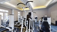 This fitness center offers a thoughtfully designed workout space outfitted with a comprehensive selection of exercise equipment. The room features multiple strength-training machines, including cable and resistance units with black-and-white framing, strategically arranged to allow comfortable movement between stations. The patterned carpet flooring adds a modern aesthetic while providing traction throughout the space.

Architecturally, the room is distinguished by a coffered ceiling with warm ambient uplighting that creates an inviting golden glow along the upper perimeter, complementing the recessed ceiling lights below. The neutral gray walls contribute to a clean, focused environment. A window along one wall introduces natural light, enhancing the overall brightness of the space.

Wall-mounted mirrors reflect the equipment and expand the visual sense of the room, making it feel open and spacious. The layout is organized to accommodate simultaneous use of multiple machines, supporting a variety of fitness routines. Overall, the fitness center balances functional design with polished finishes, creating a motivating environment well-suited for strength training and general exercise.