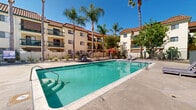 Located in Escondido, California, this outdoor pool area is a well-appointed recreational amenity set within a gated community defined by Spanish-Mediterranean architecture. The surrounding multi-story buildings feature distinctive red tile roofs, stucco exteriors, and green-railed balconies, creating a cohesive and inviting courtyard atmosphere.

At the heart of the space is a generously sized swimming pool with clear, crystal-blue water. Broad, light-toned concrete pool decking surrounds the water on all sides, providing ample room for movement and relaxation. Gentle entry steps on one end of the pool offer convenient access for swimmers of all levels.

The sundeck is furnished with multiple wood-slatted chaise lounges positioned to take advantage of Southern California's abundant sunshine. A shaded dining area featuring a table and chairs beneath a red market umbrella provides a comfortable spot for outdoor gatherings. An additional umbrella and seating arrangement can be found near a small utility structure, which houses a door providing access to facilities.

Mature palm trees and a variety of shade trees are interspersed around the perimeter, softening the hardscape and adding a lush, resort-like quality to the environment. A secured metal gate and perimeter fencing ensure privacy and safety throughout the space. The overall ambiance is bright, open, and characteristic of a relaxed Southern California outdoor lifestyle.