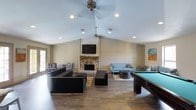 This single-floor space is designed as a versatile entertainment and lounge area, combining comfort with recreational amenities. The room's most striking architectural feature is its vaulted ceiling, which lends an expansive, open feel to the interior. Ceiling fans are mounted along the peak, ensuring good air circulation throughout the space.

The focal point of the main seating area is a stone fireplace, framed by shiplap-style wall paneling that extends upward to the ceiling ridge, giving the room a warm, rustic character. A wall-mounted flat-screen television is positioned above the fireplace, making it a natural gathering hub. Recessed lighting complements the natural brightness of the space.

The lounge area is furnished with a mix of dark modular sofas and a light blue sofa arrangement, accommodating various group sizes. Accent rugs define the seating zones atop rich, dark hardwood floors. To one side, a full-sized pool table adds a recreational dimension, perfect for social events. Natural light filters in through a side door, further enhancing the welcoming atmosphere of this well-appointed, single-level gathering space.