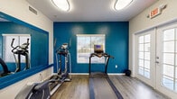This single-room fitness center offers a thoughtfully arranged workout space designed to support a full range of exercise routines. The room's most striking design element is a vivid teal accent wall, which provides a bold, energizing contrast against the surrounding neutral white walls and textured ceiling. Wood-look plank flooring runs throughout, offering both durability and a clean, modern aesthetic underfoot.

A large, blue-framed wall mirror spans one side of the room, visually expanding the space and allowing users to monitor their form during workouts. Natural light filters in through multiple windows fitted with horizontal blinds, offering outdoor views of surrounding greenery. A glass-panel entry door further contributes to the bright, open feel of the space.

The fitness room is equipped with a diverse selection of exercise equipment, including a treadmill, elliptical trainer, stationary recumbent bike, and a multi-station cable weight machine. A flat workout bench and a set of dumbbells round out the strength training options. Overhead flush-mount light fixtures provide consistent, even illumination throughout. An HVAC vent and wall-mounted thermostat ensure climate comfort during workouts. The room is accessed via a dedicated entry, making it a self-contained and functional amenity space.