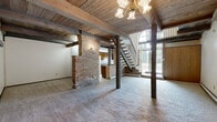 This two-floor space showcases a distinctive rustic character defined by prominent exposed wood beams running across tongue-and-groove wood plank ceilings. The main living area is generously proportioned, finished with neutral wall-to-wall carpeting and accented by vertical wood plank paneling that anchors the room with a warm, cabin-inspired aesthetic. An open railing overlooking the living space from the upper level reinforces the multi-floor layout and adds architectural interest.

The kitchen is functional and well-equipped, featuring warm-toned wood cabinetry with decorative scalloped detailing, light countertops, white appliances including a range, refrigerator, and dishwasher, and tile backsplash. A skylight above brings in natural light. The kitchen connects to the living area via a pass-through counter arrangement.

Two bathrooms serve the space. One is finished with classic beige ceramic wall tiles and features a wood vanity with a single sink and a tub area. The second bathroom, situated on the upper level beneath a sloped ceiling, features white and brown tile wainscoting, a wood vanity, and a compact, efficient layout. Both bathrooms are finished with light tile flooring.