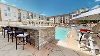 Located at 1 Richmond St in New Brunswick, NJ, this outdoor amenity space delivers a resort-style experience centered around a generously sized swimming pool with crystal-clear blue water. The pool deck is thoughtfully designed with a wide surrounding terrace finished in warm-toned pavers, accommodating an extensive arrangement of lounge chairs and umbrellas along multiple sides of the pool. A standout feature is the built-in outdoor bar, constructed with stacked natural stone and topped with a sleek counter surface, complete with bar stool seating — ideal for poolside socializing. Adjacent to the bar, a white pergola provides a shaded lounge area furnished with plush wicker sofas, accent tables, and patterned outdoor rugs, creating an inviting retreat from the sun. A raised spa or wading feature with tiled walls adds an additional amenity to the aquatic experience. The deck is framed by mature trees that provide natural greenery against the backdrop of the surrounding multi-story brick and stone residential buildings, lending the space an enclosed, community-oriented feel. Grills and additional seating areas are also present throughout the deck, making this outdoor space well-suited for both relaxation and social gatherings under open skies.