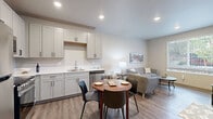 This thoughtfully designed one-bedroom, one-bathroom unit offers an efficient and comfortable layout across a single floor. The open-concept kitchen and living area serves as the heart of the unit, featuring light gray shaker-style cabinetry, white countertops, and a full suite of stainless steel appliances including a refrigerator, range, microwave, and dishwasher. Recessed lighting illuminates the space, while durable wood-look flooring ties the kitchen, dining, and living areas together seamlessly. A round dining table nestled between the kitchen and living room provides a natural transition between the two spaces. The living area is bright and inviting, benefiting from a large window that frames outdoor views and fills the room with natural light. The bedroom is carpeted for comfort, features a generous sliding-door closet, and includes a sizable window overlooking greenery outside. The bathroom is equipped with a single-sink vanity, wood-look flooring, and accessibility-conscious grab bars. The unit also includes a dedicated laundry area, adding everyday convenience. Neutral tones and clean lines throughout create a modern, cohesive aesthetic.