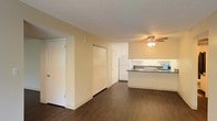 This single-floor space showcases an open-concept layout that seamlessly connects the living area to the kitchen, creating a functional and cohesive environment. The dark hardwood-style flooring runs throughout, providing a warm contrast to the light beige walls and white trim. A textured ceiling adds subtle dimension to the interior.

The living area is generously proportioned, offering ample room for furniture arrangement. Multiple doors along the back wall suggest convenient closet or storage access, enhancing the unit's practical appeal. A ceiling fan with an integrated light fixture is centrally positioned, providing both air circulation and ambient lighting to the space.

The kitchen is positioned adjacent to the living area and features white cabinetry that extends to the ceiling for maximum storage. A white refrigerator anchors one end of the kitchen, while a peninsula with a darker countertop defines the boundary between the two spaces. A sink with a window or backsplash detail adds a finishing touch. The overall palette of whites, neutrals, and warm wood tones gives the space a clean, approachable aesthetic.