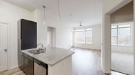 Located at 1100 South Boulevard in Charlotte's vibrant South End neighborhood, this two-bedroom, two-bathroom residence offers a well-designed single-floor layout with standout architectural details throughout. The open-concept living, dining, and kitchen area is anchored by impressively high ceilings that amplify the sense of space. The kitchen features a curved granite-topped peninsula with an undermount sink, dark espresso cabinetry, stainless steel appliances including a refrigerator and range, subway tile backsplash, and stylish pendant lighting. Wood-look plank flooring runs throughout the main living areas, complementing the clean, neutral color palette. Large windows in the living room and primary bedroom frame city skyline views and flood the interior with natural light, each accented by ceiling fans. The primary bedroom includes an en-suite bathroom with dark vanity cabinetry, granite countertops, and a glass-enclosed shower. The secondary bedroom features plush carpeting and its own city-facing window. Three closets provide ample storage, including a walk-in closet with wire shelving. A dedicated laundry area with an in-unit washer adds everyday convenience. The hallway connects all rooms efficiently, completing this thoughtfully laid-out urban residence.
