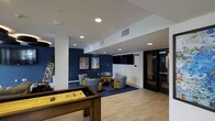 This single-floor amenity lounge is thoughtfully designed to offer both entertainment and relaxation in a contemporary setting. The space is defined by a clean, modern aesthetic, featuring a crisp white ceiling with recessed lighting that evenly illuminates the room, complemented by light hardwood-style flooring that adds warmth throughout.

A standout feature is the bold navy blue accent wall, which anchors a cozy seating area furnished with stylish patterned lounge chairs and coordinating yellow accent pieces arranged around a central coffee table. Abstract artwork adorns the wall, adding a creative touch, while a large flat-screen television serves as a focal point for entertainment.

Adjacent to the lounge seating, a shuffleboard table takes center stage, providing a dedicated game zone that invites social interaction and recreational play. The open layout ensures easy flow between the gaming and lounge areas.

Structural columns are integrated seamlessly into the design, maintaining the open feel of the space. A glass-panel door is visible along one wall, offering a sense of transparency and connectivity. The overall ambiance is polished yet inviting, making this lounge an ideal communal space for socializing, gaming, and unwinding.