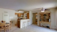 This single-floor space is designed with an open-concept layout that seamlessly connects the kitchen and living area. The kitchen features warm honey-toned wooden cabinetry, a gas range, and under-cabinet lighting, creating a practical yet inviting cooking space. A peninsula with a raised breakfast bar extends from the kitchen, accommodating three bar stools and serving as a natural divider between the kitchen and the living area. The living area is finished with plush wall-to-wall carpeting, contributing to a cozy atmosphere, while a ceiling fan with an integrated light fixture provides both comfort and illumination. A freestanding wooden shelving unit with drawers adds storage and display space along one wall. The textured ceiling is consistent throughout, adding subtle visual interest. A hallway visible from the main living area leads to additional rooms, hinting at a well-organized floor plan. Neutral beige tones dominate the walls and flooring, creating a cohesive and calming aesthetic throughout the space.