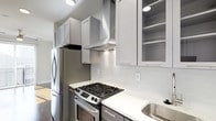 The kitchen is thoughtfully designed with a clean, modern aesthetic that balances form and function. Gray shaker-style cabinetry lines the walls, extending floor-to-ceiling to maximize storage capacity. Upper cabinets include glass-front panels on select units, offering an opportunity to display dishware while maintaining a cohesive look. The crisp white countertops provide a bright contrast against the gray cabinetry, creating a visually striking workspace. A classic white subway tile backsplash adds texture and a timeless touch to the cooking area. Stainless steel appliances, including a gas range with multiple burners, a built-in oven, and a full-size refrigerator, outfit the kitchen with professional-grade tools. A stainless steel range hood is mounted above the cooktop, ensuring proper ventilation. Recessed lighting installed in the ceiling provides focused illumination throughout the space. The stainless steel sink is positioned conveniently along the countertop. Dark hardwood flooring grounds the space and adds warmth, complementing the cooler tones of the cabinetry and appliances. Overall, the kitchen presents a polished and highly functional environment.