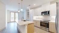 This thoughtfully designed 1-bedroom, 1-bathroom unit offers a bright, modern living experience across a single floor. The open-concept layout centers around a well-appointed kitchen featuring a large quartz-topped center island with an undermount sink and gold-accented faucet. Upper white cabinetry with brass hardware pairs with warm wood-tone lower cabinets, complemented by a soft subway tile backsplash. Stainless steel appliances — including a French door refrigerator, electric range, and over-the-range microwave — complete the space. Pendant globe lights with brass fixtures add an elegant touch overhead. Light hardwood-style flooring flows throughout, unifying the kitchen, living area, and beyond. Built-in open shelving provides additional storage near the living space, which also includes an in-unit washer and dryer. The living area benefits from natural light and ceiling fan. The bedroom is a serene, well-proportioned retreat with a ceiling fan, natural light from a window, and access to a private balcony via French doors. A walk-in closet with built-in shelving and hanging rods serves the bedroom. The bathroom features a glass-enclosed walk-in shower with floor-to-ceiling dark tile and a built-in bench, finished with sleek gold hardware.