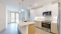 This thoughtfully designed 1-bedroom, 1-bathroom unit offers a bright, modern living experience across a single floor. The open-concept layout centers around a well-appointed kitchen featuring a large quartz-topped center island with an undermount sink and gold-accented faucet. Upper white cabinetry with brass hardware pairs with warm wood-tone lower cabinets, complemented by a soft subway tile backsplash. Stainless steel appliances — including a French door refrigerator, electric range, and over-the-range microwave — complete the space. Pendant globe lights with brass fixtures add an elegant touch overhead. Light hardwood-style flooring flows throughout, unifying the kitchen, living area, and beyond. Built-in open shelving provides additional storage near the living space, which also includes an in-unit washer and dryer. The living area benefits from natural light and ceiling fan. The bedroom is a serene, well-proportioned retreat with a ceiling fan, natural light from a window, and access to a private balcony via French doors. A walk-in closet with built-in shelving and hanging rods serves the bedroom. The bathroom features a glass-enclosed walk-in shower with floor-to-ceiling dark tile and a built-in bench, finished with sleek gold hardware.
