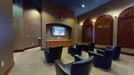 This single-floor media lounge is thoughtfully designed to deliver a premium viewing and social experience. At the focal point of the room, a large flat-screen display is mounted within a rounded archway alcove, flanked by in-wall speakers and supported by a row of warm-toned wooden cabinetry below, providing both style and functional storage. The seating arrangement consists of numerous sleek black leather swivel chairs with built-in cup holders, complemented by matching leather ottomans, all oriented toward the screen for optimal viewing. The room's color palette pairs rich, earthy brown walls with a deep navy-blue ceiling, creating a cozy, immersive atmosphere. A standout architectural feature is the row of tall, arched wood-shuttered windows lining the side wall, their dark mahogany finish adding a classic, sophisticated character to the space. Wall sconces with warm-toned bulbs are positioned throughout, casting a soft, ambient glow that enhances the lounge's inviting feel. Neutral patterned carpeting runs throughout the main viewing area, while tiled flooring marks the entryway transition. A decorative acoustic panel is suspended from the ceiling above, contributing to both the room's aesthetics and sound quality. An adjacent area, visible through a wood-trimmed doorway, hints at additional amenities nearby.