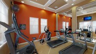 This fitness center is designed to provide a comprehensive cardio-focused workout experience within a single-floor space. The room immediately draws attention with its bold, burnt-orange accent walls contrasted against warm tan walls, creating an energizing atmosphere conducive to exercise. The ceiling features elegant coffered detailing with recessed lighting that evenly illuminates the space throughout.

The cardio equipment selection is well-rounded, including multiple treadmills, elliptical trainers, an upright stationary bike, and a recumbent-style machine, accommodating a variety of workout preferences. Large framed mirrors line one wall, visually expanding the room and allowing users to monitor their form during exercise. A wall-mounted flat-screen television is positioned for easy viewing, accompanied by two wall-mounted fans that ensure comfortable air circulation.

Natural light filters in through several windows fitted with white horizontal blinds, adding brightness while maintaining privacy. A built-in shelving unit with orange-painted interior provides storage for fitness accessories or towels. A water dispenser station is conveniently positioned nearby. A glass-paneled door leads to what appears to be an outdoor area, further enhancing ventilation and accessibility. The carpeted flooring completes the space with a practical, comfortable surface underfoot.