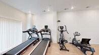 Located at 1310 E St. Louis Street in Springfield, Missouri, this fitness center offers a clean, modern environment designed to support a full range of workout routines. The space is laid out across a single level and makes efficient use of its compact footprint.

The exercise room features light hardwood-style flooring throughout, crisp white walls, and recessed ceiling lighting that keeps the space bright and energizing. A large floor-to-ceiling mirror spans one accent wall, finished in a warm taupe tone, which visually expands the room and assists users during strength training. Natural light filters in through windows equipped with vertical blinds, adding to the welcoming atmosphere.

Cardio equipment includes multiple treadmills and elliptical trainers, along with a stationary recumbent bike, providing users with a variety of aerobic options. A dedicated free weights area features an adjustable dumbbell rack and a workout bench, complemented by a rubber mat for floor exercises. An additional open area along one wall offers extra space for stretching or functional training.

A conveniently located bathroom adjoins the exercise room, featuring light wood-look flooring, a wall-mounted sink with a framed mirror, accessibility grab bars, and a clean, minimalist design. The overall ambiance of the fitness center is modern, functional, and well-maintained — well-suited for residents or users seeking a convenient on-site workout facility.