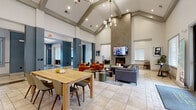 Located at 120 Cinema Dr in Hendersonville, Tennessee, this single-story community clubhouse is designed to offer a sophisticated and welcoming environment for residents. The centerpiece of the space is a grand open lounge area distinguished by dramatic vaulted ceilings with exposed beam detailing and a striking floor-to-ceiling stone fireplace topped with a wall-mounted flat-screen TV. Comfortable seating arrangements — including plush gray sofas and warm terracotta accent chairs — are thoughtfully positioned on patterned area rugs over light tile flooring, creating an inviting atmosphere for socializing or relaxing. Natural light pours in through multiple large windows dressed with horizontal blinds, with views overlooking an outdoor pool area. The fully equipped communal kitchen features sage-green raised-panel cabinetry, granite countertops, a tile backsplash, a central island with bar seating, and full-size appliances including a black French door refrigerator and wall oven. A dedicated business center offers a large conference table, computer workstations, a printer, a wall-mounted display, and a quiet lounge nook with upholstered seating — ideal for remote work or small meetings. Throughout the space, a cohesive modern aesthetic is maintained with neutral tones, contemporary furnishings, and carefully chosen artwork.