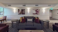This thoughtfully designed single-floor communal space combines elegance and functionality across several interconnected areas. At its heart is a generously sized lounge featuring a striking white brick fireplace with a wall-mounted television above, flanked by sculptural sconce lighting and lush potted greenery. The fireplace serves as a natural focal point, anchoring the seating area furnished with plush gray sofas and deep burgundy armchairs accented by mustard and crimson throw pillows.

A series of boldly colored abstract artworks in pink, burgundy, and earth tones line the walls, complementing the refined yet welcoming atmosphere. Wall sconces throughout provide warm, ambient lighting, while large windows along one side of the lounge invite natural light and offer views of mature trees outside.

The open-plan layout transitions seamlessly into a kitchen-style service area featuring an expansive white marble countertop with subtle gray veining, a matte black sink and faucet, and bar-height seating. Adjacent to this is a dedicated refreshment corridor outfitted with dark cabinetry, light tile flooring, stainless steel appliances, a coffee station, and vibrant horizontal-gradient artwork decorating the walls. The overall ambiance is polished and contemporary, designed to encourage relaxed social interaction.