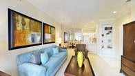 This single-floor residence offers a thoughtfully designed open-concept layout that seamlessly connects the living, dining, and kitchen areas into one cohesive and functional space. The living area serves as the focal point, showcasing plush blue seating, bold abstract wall art, and a dark wood coffee table that anchors the room with contrast and character. Warm neutral tones on the walls and light carpeting create a welcoming ambiance throughout. Recessed lighting is strategically placed across the ceiling, providing bright, even illumination that enhances the sense of openness. A stylish floor lamp adds a softer, layered lighting element to the seating area. Beyond the living space, a compact dining area with a round table and chairs sits just before the kitchen, which features a pass-through counter and white cabinetry — an efficient and accessible design ideal for entertaining. The kitchen is well-lit and neatly appointed, with clean lines that complement the overall aesthetic. Built-in shelving is visible along one wall, offering additional display and storage options. The entire space is unified by its consistent warm palette, modern finishes, and a balance of comfort and style that makes it both practical and inviting.