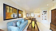 This single-floor residence offers a thoughtfully designed open-concept layout that seamlessly connects the living, dining, and kitchen areas into one cohesive and functional space. The living area serves as the focal point, showcasing plush blue seating, bold abstract wall art, and a dark wood coffee table that anchors the room with contrast and character. Warm neutral tones on the walls and light carpeting create a welcoming ambiance throughout. Recessed lighting is strategically placed across the ceiling, providing bright, even illumination that enhances the sense of openness. A stylish floor lamp adds a softer, layered lighting element to the seating area. Beyond the living space, a compact dining area with a round table and chairs sits just before the kitchen, which features a pass-through counter and white cabinetry — an efficient and accessible design ideal for entertaining. The kitchen is well-lit and neatly appointed, with clean lines that complement the overall aesthetic. Built-in shelving is visible along one wall, offering additional display and storage options. The entire space is unified by its consistent warm palette, modern finishes, and a balance of comfort and style that makes it both practical and inviting.