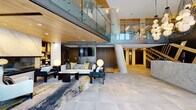 This striking lobby space makes an immediate impression with its soaring double-height ceilings and floor-to-ceiling glazed entry doors that flood the interior with natural light and offer clear sightlines to the surrounding streetscape. Large-format stone-look floor tiles extend throughout, grounding the space with a sense of refined permanence.

At the heart of the lobby stands a substantial marble reception desk, positioned beneath a dramatic cluster of globe pendant chandeliers. Behind it, a bold geometric feature wall rendered in contrasting dark and gold tones adds artistic depth and visual energy. A floating staircase with open wood treads and sleek glass railings rises elegantly to an upper mezzanine level, reinforcing the vertical grandeur of the space.

A dedicated lounge area offers a welcoming retreat, anchored by a plush white sofa accented with chartreuse and blue cushions, a marble-topped coffee table, and a built-in fireplace framed by curated open shelving. Warm wood ceiling panels and soft ambient lighting create an inviting contrast to the polished stone surfaces.

The elevator lobby is thoughtfully finished with natural wood wall paneling, marble tile accents, navy elevator doors, and integrated digital directories—demonstrating consistent attention to material quality and detail throughout.