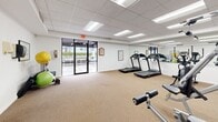 Located at 2903–2915 NW 60th Ave in Sunrise, Florida, this single-floor fitness center offers a well-equipped workout space designed to support a variety of exercise routines. The room features a drop-panel ceiling with recessed fluorescent lighting that keeps the space bright and functional throughout. Neutral-toned carpet covers the floor, providing a comfortable surface underfoot across the entire workout area.

The cardio section is prominently equipped with multiple treadmills and an elliptical trainer, arranged in a row along one side of the room. A full-wall mirror spans this area, enhancing the sense of openness and allowing users to monitor their form during workouts. On the opposite side, a multi-station cable and weight machine anchors the strength training area, accompanied by a stationary recumbent bike and a leg extension/curl bench.

A dedicated accessory corner houses stability balls, medicine balls, resistance tools, and a fitness information rack, offering additional variety for functional training. The space is finished with motivational artwork displayed on the white walls, adding an energetic ambiance. A glass double-door entry provides direct access to an outdoor area, allowing natural light to filter in and contributing to an open, airy feel. The overall layout is practical and efficiently organized for a comprehensive fitness experience.