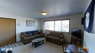 This single-floor space offers a warm and functional layout designed for everyday living. The living room serves as the central gathering area, featuring wall-to-wall carpeting that adds a sense of warmth and comfort underfoot. Two generously sized sofas in earthy brown tones are arranged around an oval coffee table, creating an inviting seating arrangement ideal for relaxing or entertaining. A large multi-pane picture window dominates one wall, allowing an abundance of natural light to fill the room and providing views of the surrounding exterior. A flat-screen television is positioned on the opposite side of the room, complemented by a wooden side table and chair that add a classic touch. The ceiling features a textured popcorn finish with a centrally mounted flush light fixture that provides ambient illumination. The neutral-toned walls keep the space feeling open and adaptable. Wall art adds a personal touch to the room, contributing to the overall lived-in and welcoming atmosphere. The space is straightforward and practical, offering a comfortable, no-frills environment suited for daily use.