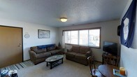 This single-floor space offers a warm and functional layout designed for everyday living. The living room serves as the central gathering area, featuring wall-to-wall carpeting that adds a sense of warmth and comfort underfoot. Two generously sized sofas in earthy brown tones are arranged around an oval coffee table, creating an inviting seating arrangement ideal for relaxing or entertaining. A large multi-pane picture window dominates one wall, allowing an abundance of natural light to fill the room and providing views of the surrounding exterior. A flat-screen television is positioned on the opposite side of the room, complemented by a wooden side table and chair that add a classic touch. The ceiling features a textured popcorn finish with a centrally mounted flush light fixture that provides ambient illumination. The neutral-toned walls keep the space feeling open and adaptable. Wall art adds a personal touch to the room, contributing to the overall lived-in and welcoming atmosphere. The space is straightforward and practical, offering a comfortable, no-frills environment suited for daily use.